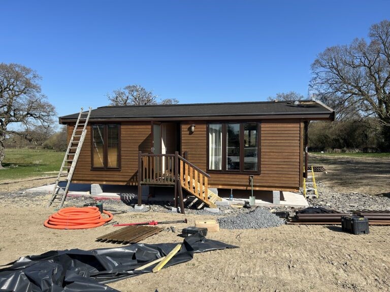 a Pinelog timber lodge is being constructed on a site. there is a set of step ladders against thd lodge and a man is on the roof working. on the ground there are building materials, including a coil of bright orange piping, black plastic sheeting