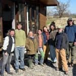 A group of male and female people smiling and wearing outdoor clothes standing outside the entrance to a Pinelog timber lodge. There is blue sky and there are fields and trees in the background